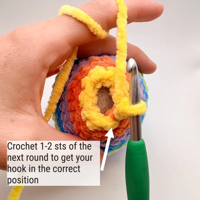 A close-up of the back of a crocheted fish's tail with a green crochet hook inserted in a stitch. Text on the image reads "Crochet 1-2 sts of the next round to get your hook in the correct position," with a white arrow pointing to a stitch on the tail.