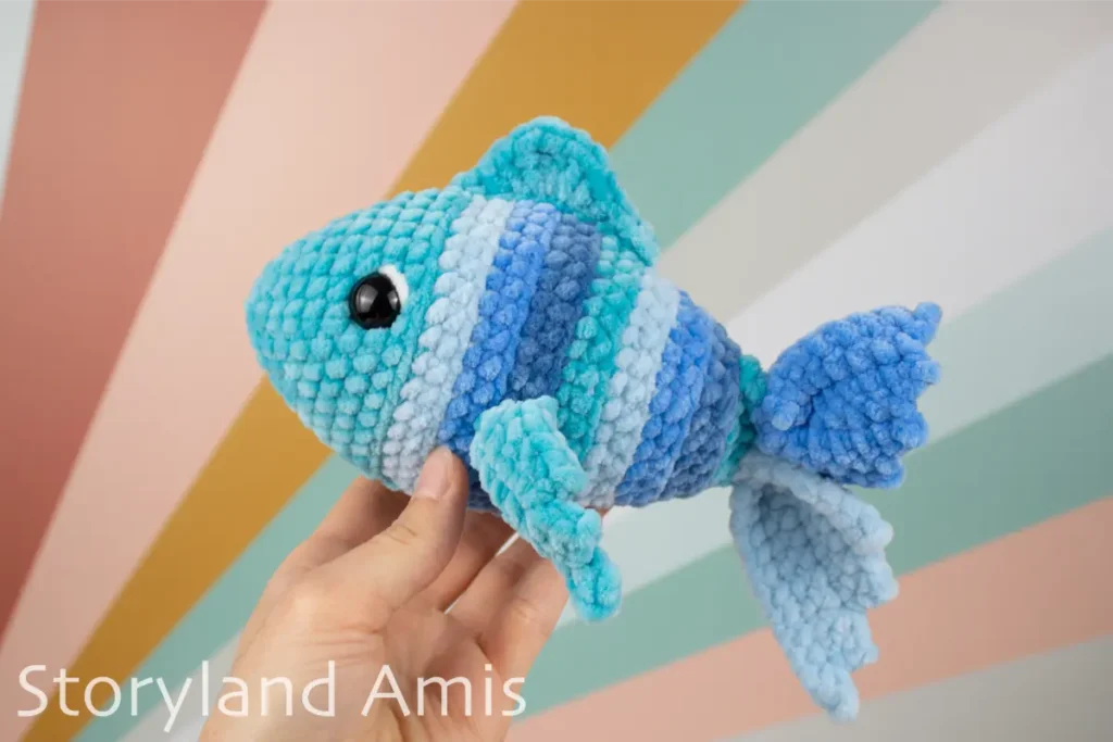 A hand holds up a blue, crocheted fish against a colorful backdrop. 