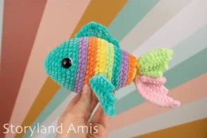 A hand holds up a crocheted multicolored fish in front of a colorful background.