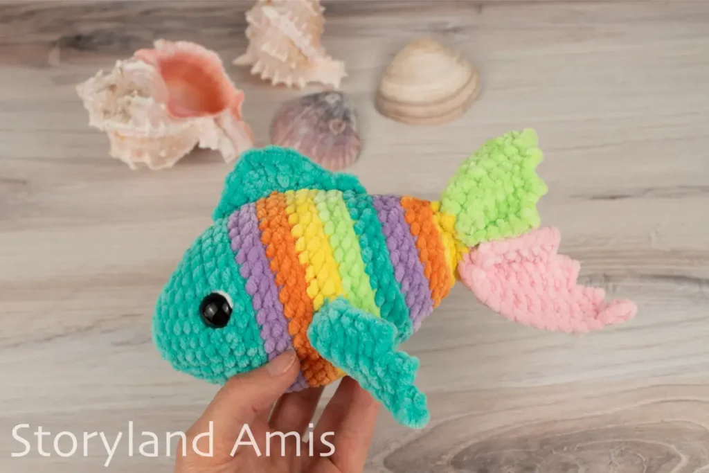 A hand holds a multicolored amigurumi fish against a wooden table with seashells in the background. 
