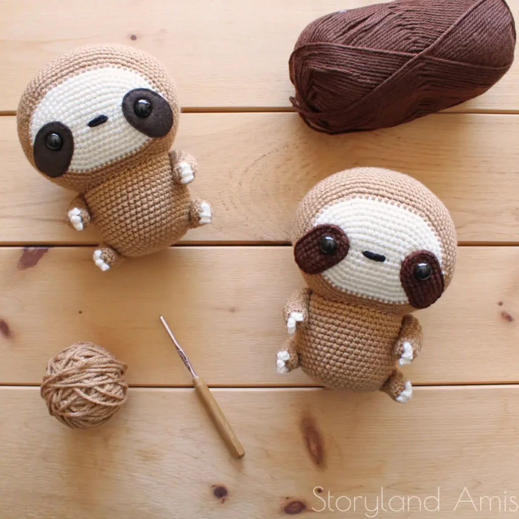Two crocheted sloths lay on a wooden table. A crochet hook and two balls of yarn lie around them.