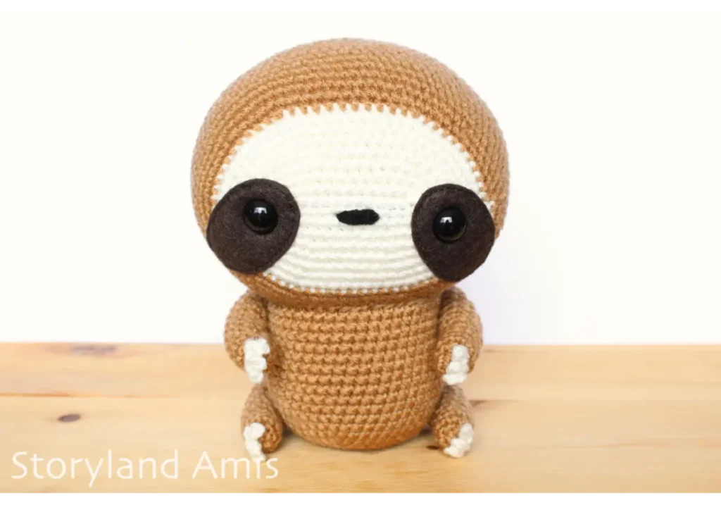 A crocheted amigurumi sloth sits on a wooden table.