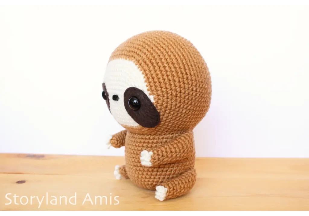 A crocheted amigurumi sloth sits on a wooden table.