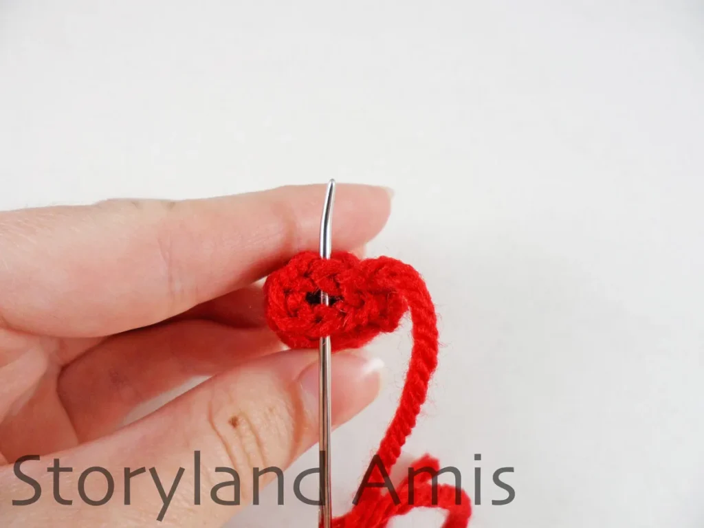 A hand holds a small crocheted doll arm with a tapestry needle going through the stitches on the last round of the arm.