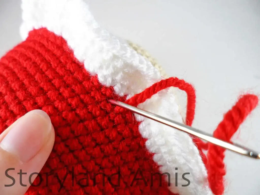 A close-up of a tapestry needle being used to sew a red and white crocheted cap to an amigurumi Mrs. Claus's head.