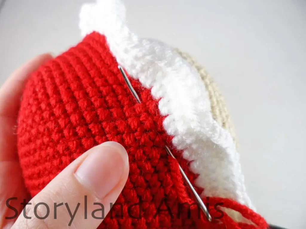 A close-up of a tapestry needle being used to sew a red and white crocheted cap to an amigurumi Mrs. Claus's head.