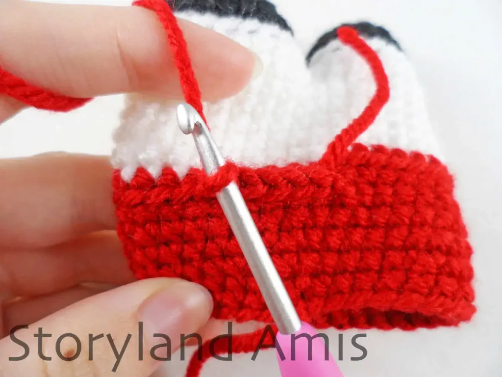 A close-up of a crochet hook inserted into a stitch of a crocheted amigurumi doll body. A hand holds the body steady.
