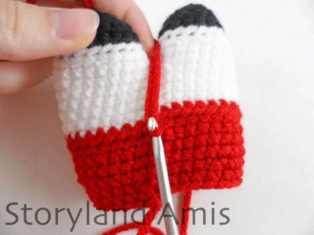 A close-up of a crochet hook inserted into a stitch of a crocheted amigurumi doll body. A hand holds the body steady.