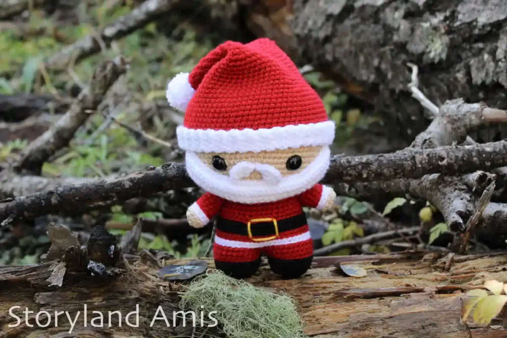 A crocheted amigurumi Santa Claus doll stands in a forest with sticks and moss around it.