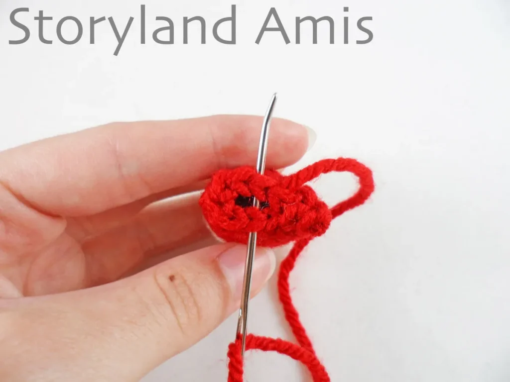 A hand holds a small crocheted doll arm with a tapestry needle going through the stitches on the last round of the arm.