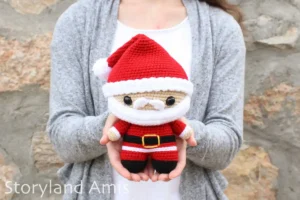 A woman wearing a white shirt and a gray sweater stands in front of a rock wall holding a crocheted amigurumi Santa Claus doll.