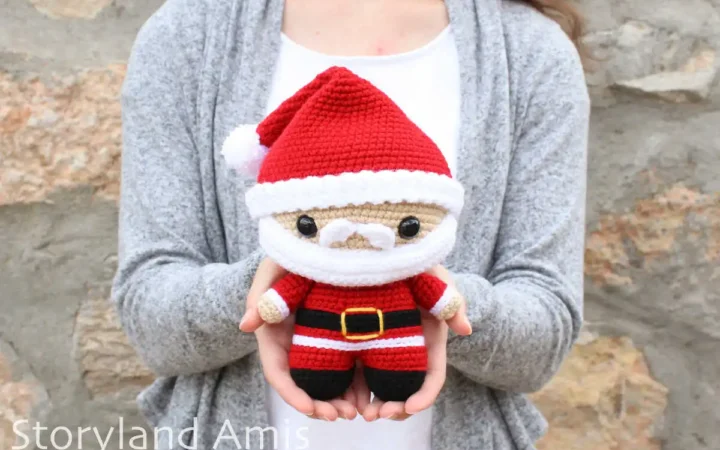 A woman wearing a white shirt and a gray sweater stands in front of a rock wall holding a crocheted amigurumi Santa Claus doll.