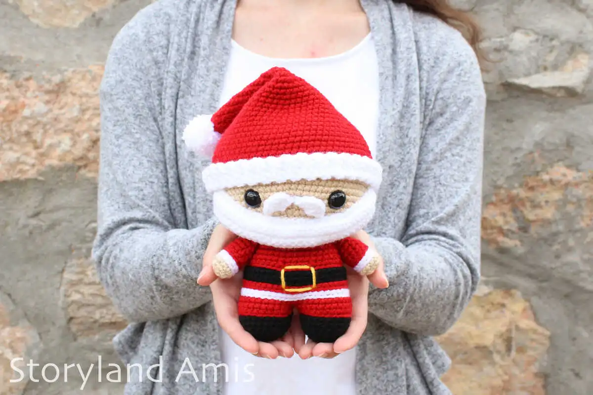 A woman wearing a white shirt and a gray sweater stands in front of a rock wall holding a crocheted amigurumi Santa Claus doll.