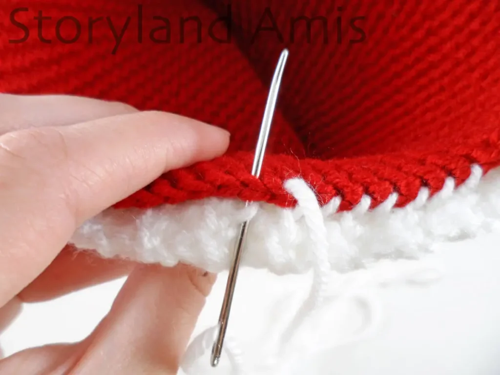 A close-up of a hand using a tapestry needle to sew a white crocheted band to the edge of a red hat.