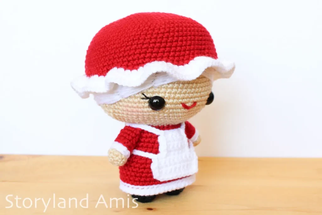 A crocheted Mrs. Claus amigurumi doll stands on a wooden table against a white background.