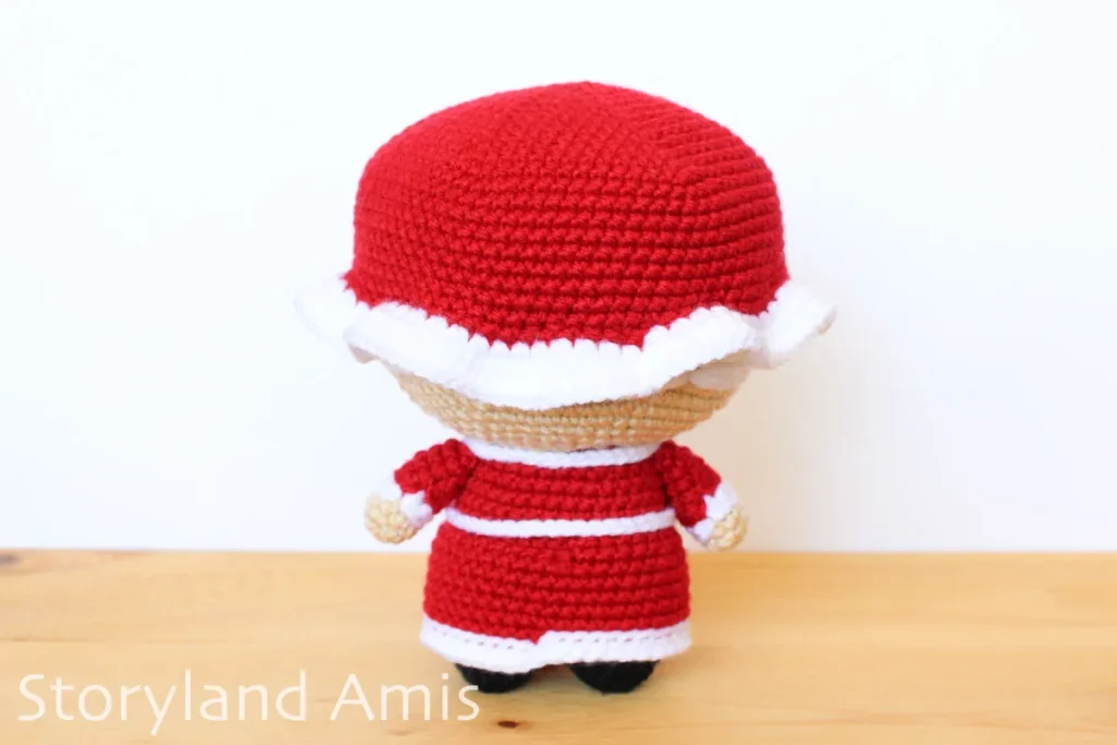 A crocheted Mrs. Claus amigurumi doll stands on a wooden table against a white background.