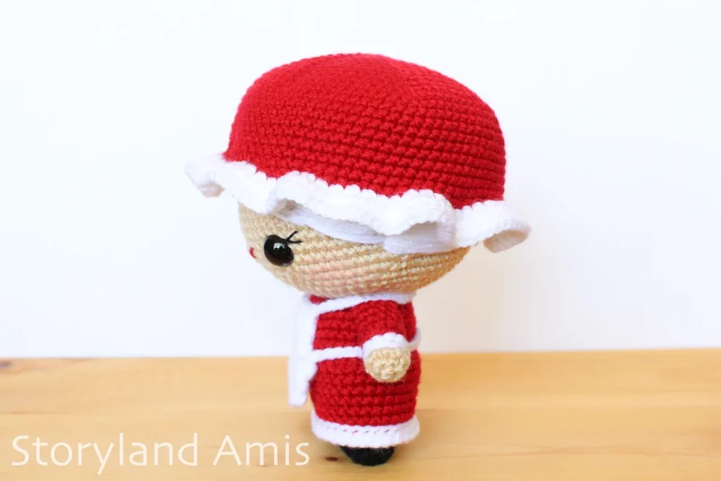 A crocheted Mrs. Claus amigurumi doll stands on a wooden table against a white background.