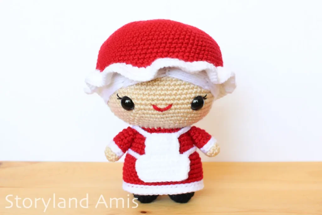 A crocheted Mrs. Claus amigurumi doll stands on a wooden table against a white background.