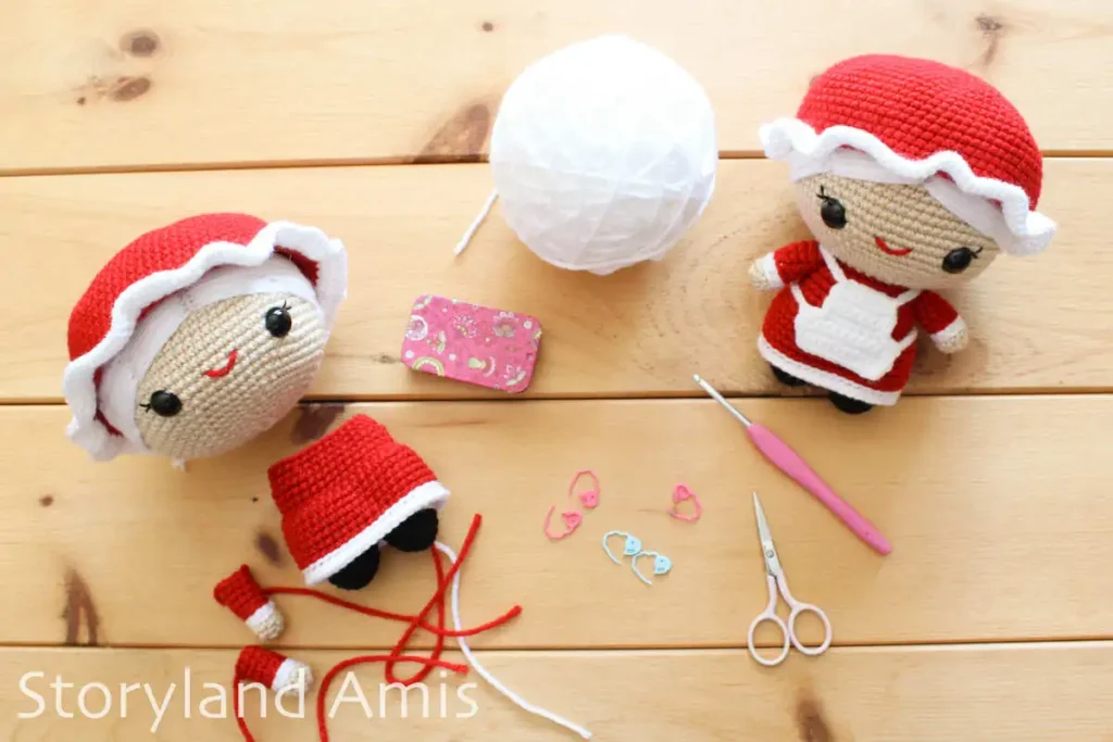 A partially completed amigurumi Mrs. Claus doll lays on a wooden table with a completed Mrs. Claus doll close by. Yarn, stitch markers, scissors, and a crochet hook lay scattered over the table between the two dolls.