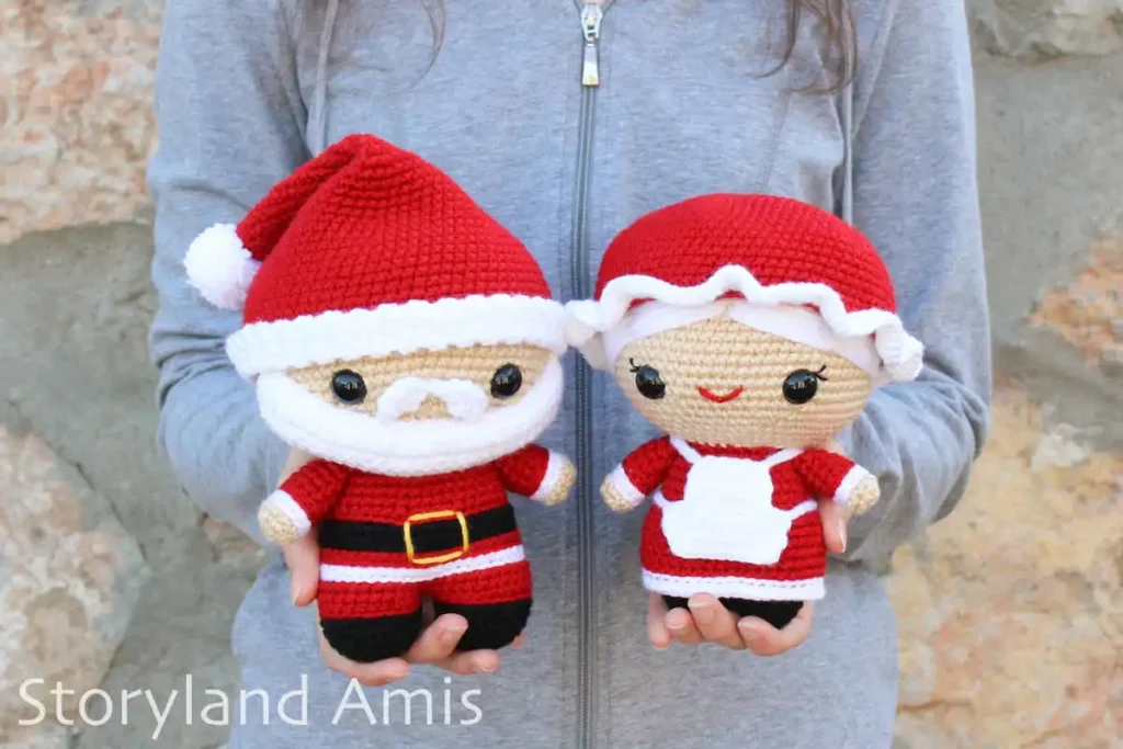 A girl wearing a gray hoodie holds a crocheted amigurumi Santa Claus and Mrs. Claus dolls while standing in front of a rock wall.