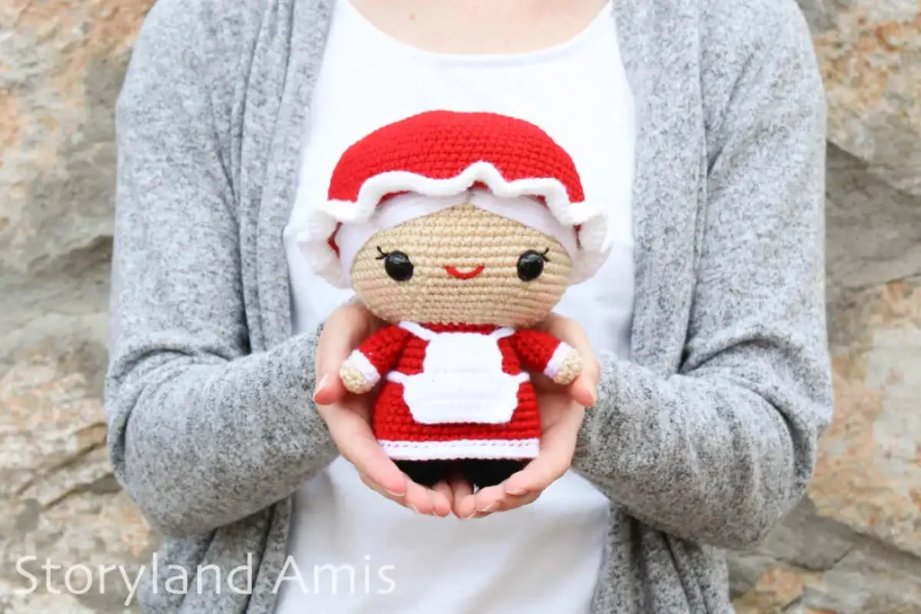 A girl wearing a white shirt and a gray sweater holds a crocheted amigurumi Mrs. Claus doll while standing in front of a rock wall.