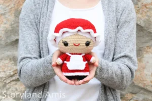 A girl wearing a white shirt and a gray sweater holds a crocheted amigurumi Mrs. Claus doll while standing in front of a rock wall.