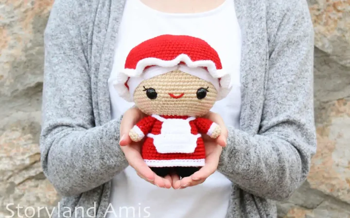 A girl wearing a white shirt and a gray sweater holds a crocheted amigurumi Mrs. Claus doll while standing in front of a rock wall.