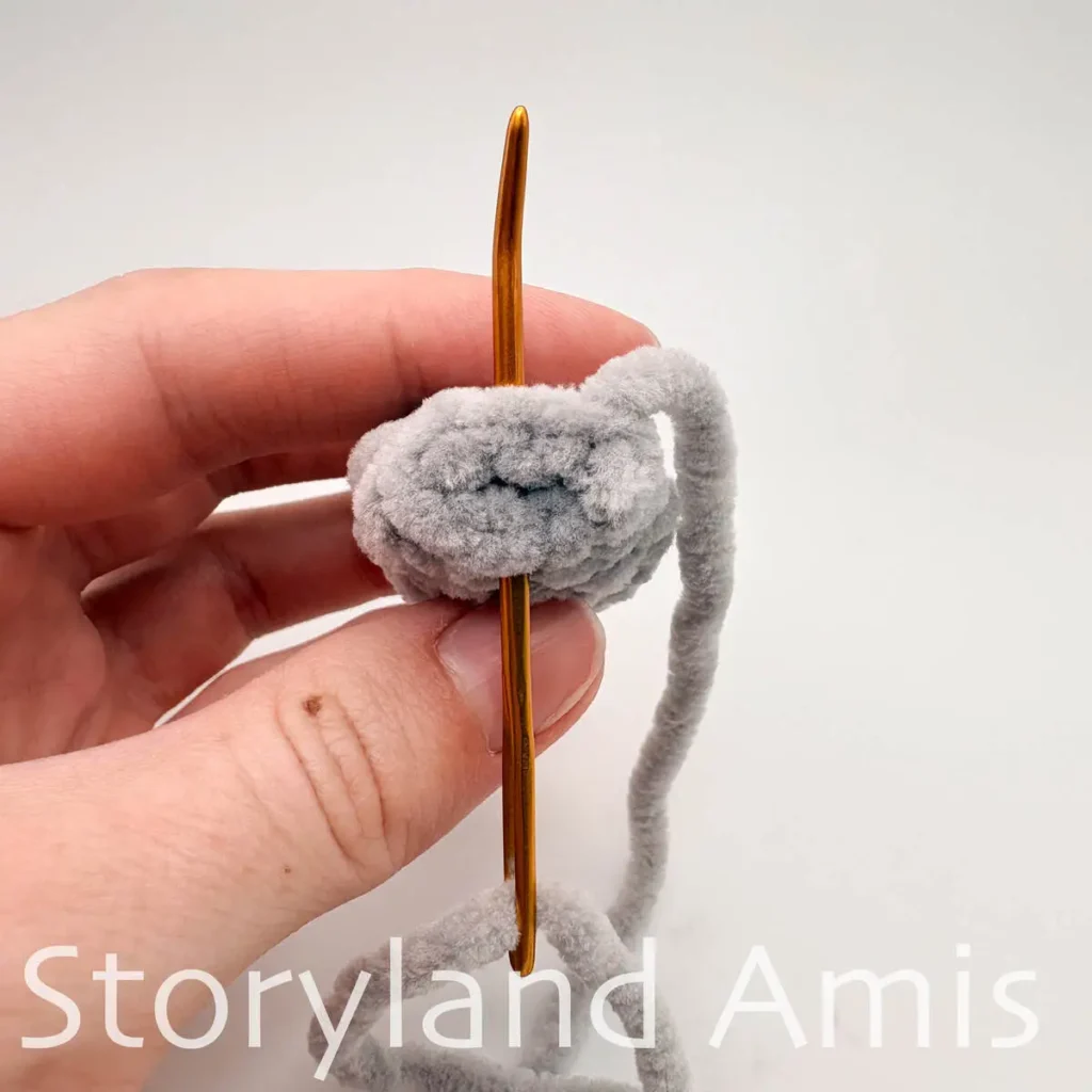 A hand holds a gray crocheted manatee flipper with a tapestry needle going through the stitches on the last round of the flipper.