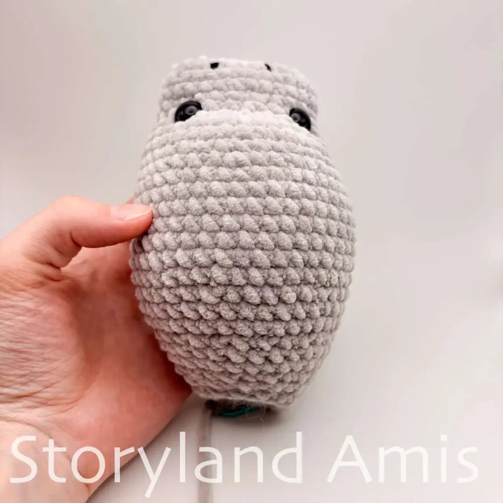 A hand holds up a partially finished amigurumi manatee plush toy. The toy is shown from above.