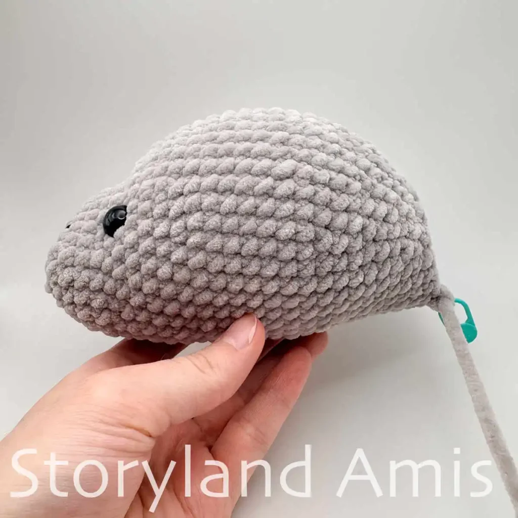 A hand holds up a partially finished amigurumi manatee plush toy. The toy is shown from the side.