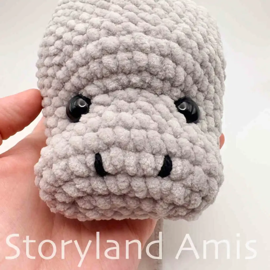 A close-up of the face of a partially finished amigurumi manatee with black embroidered nostrils and black plastic safety eyes.