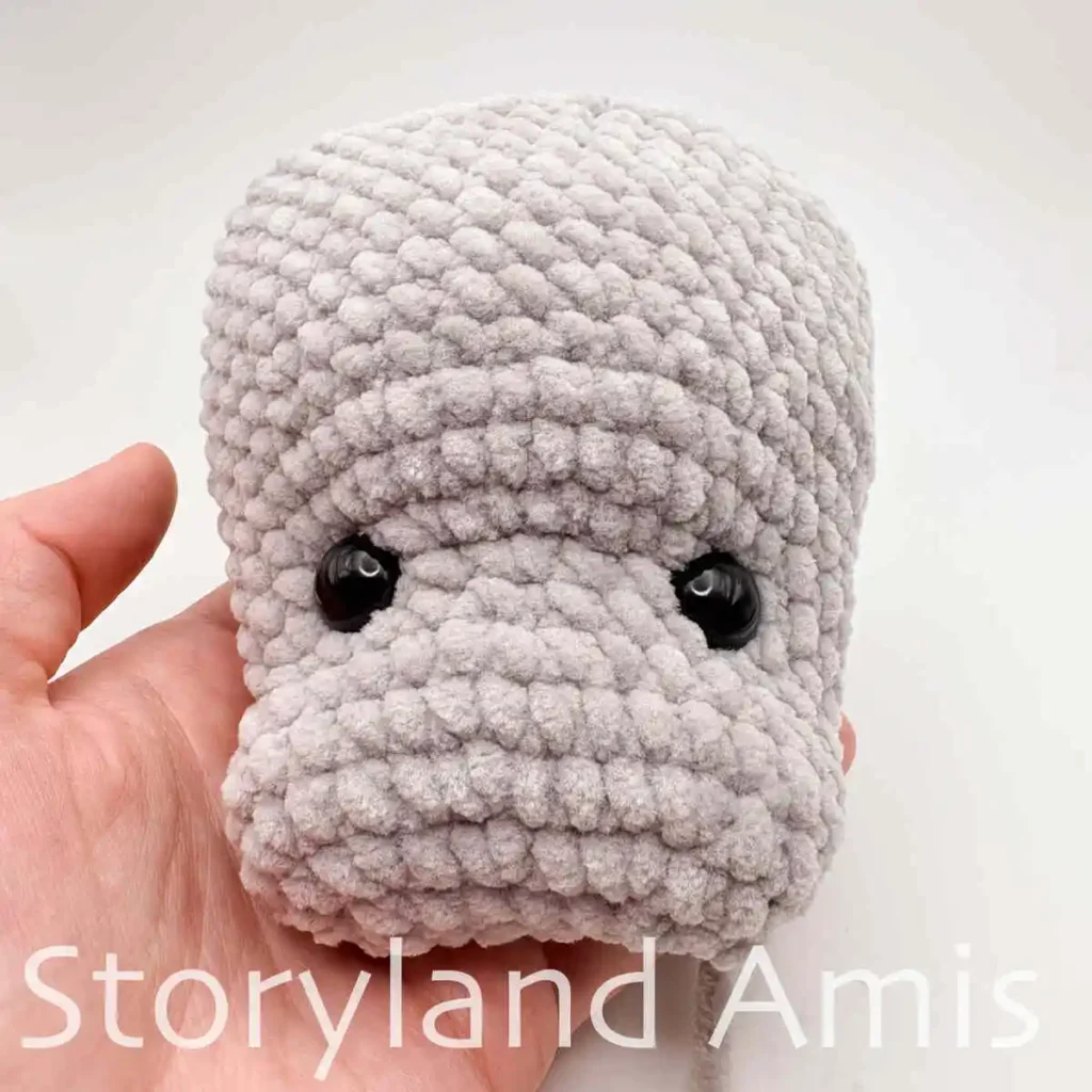 A hand holds a partially finished amigurumi manatee with two black plastic safety eyes inserted into the head.
