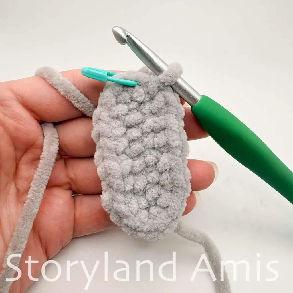 A hand holds a gray crocheted oval piece with a green crochet hook inserted into the corner stitch next to a blue stitch marker.