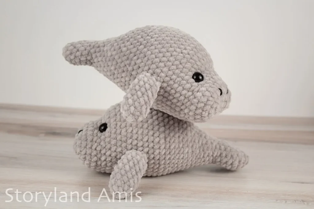 Two crocheted manatee amigurumi toys stacked on top of each other sit on a wooden table.