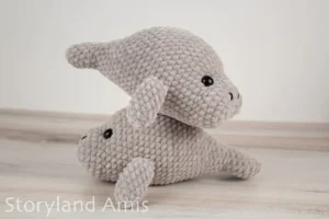 Two crocheted manatee amigurumi toys stacked on top of each other sit on a wooden table.