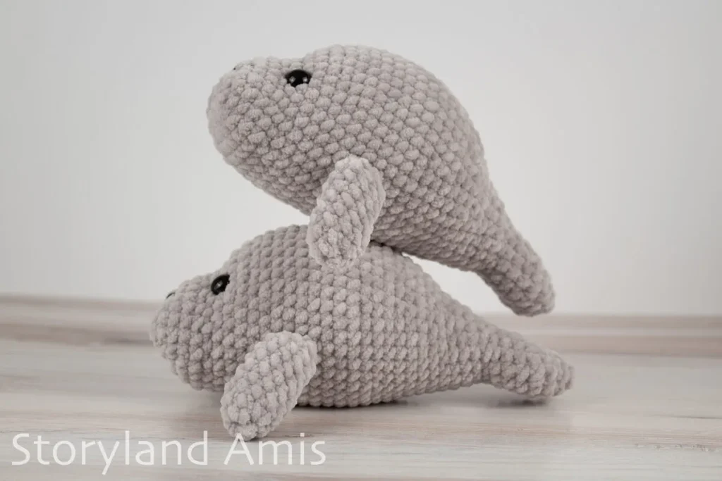 Two crocheted manatee amigurumi toys stacked on top of each other sit on a wooden table.