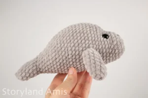 A hand holds a crocheted amigurumi manatee toy up against a white background.