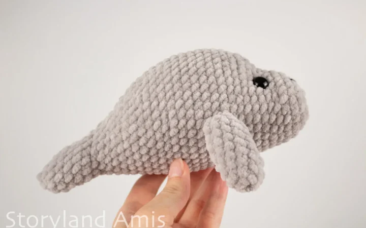 A hand holds a crocheted amigurumi manatee toy up against a white background.