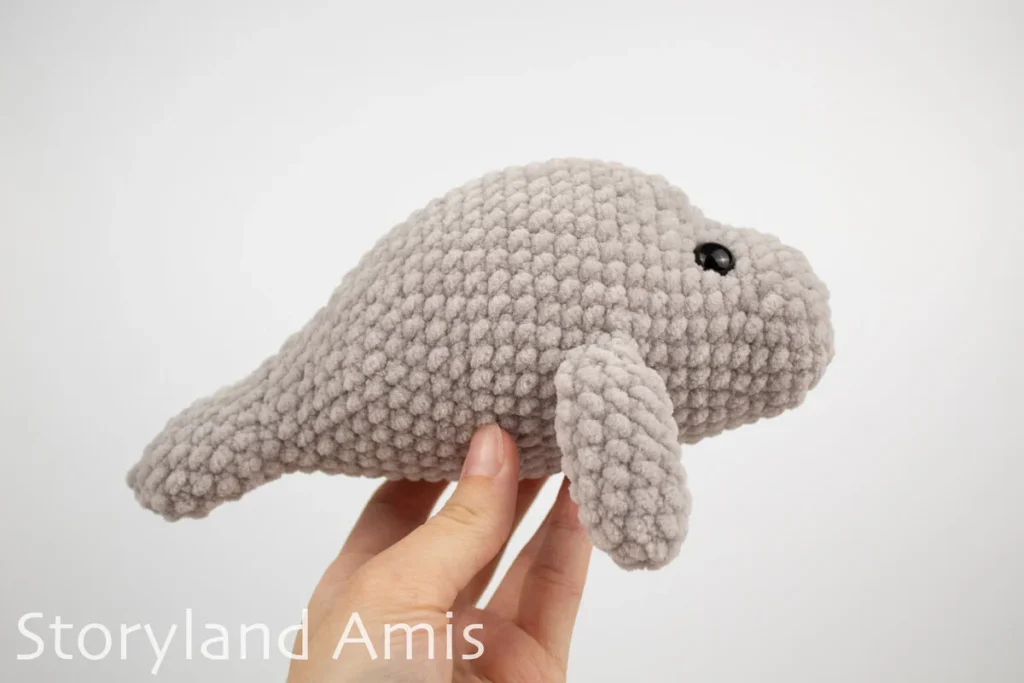 A hand holds a crocheted amigurumi manatee toy up against a white background.