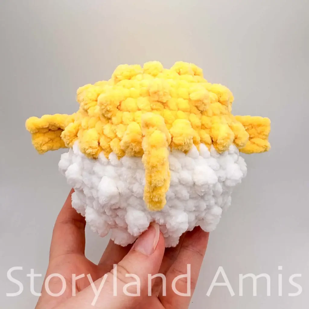 A hand holds an amigurumi pufferfish. The fish is shown from the back.