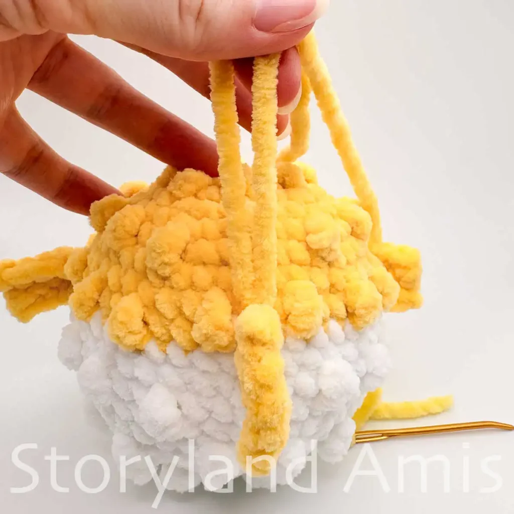 A close-up of the back of an amigurumi pufferfish. A hand holds two yellow yarn tails that are coming out of the fish's tail.