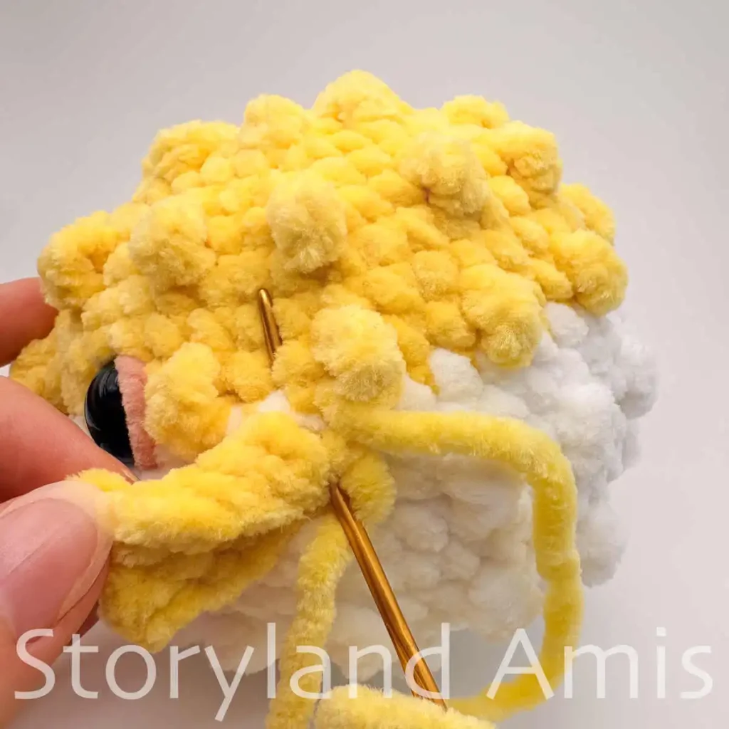 A close-up of a hand using a tapestry needle to sew a crocheted fin to the body of an amigurumi pufferfish.