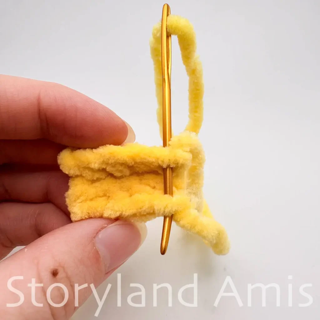 A hand holds a small yellow crocheted rectangle with a tapestry needle inserted in the edge stitches.