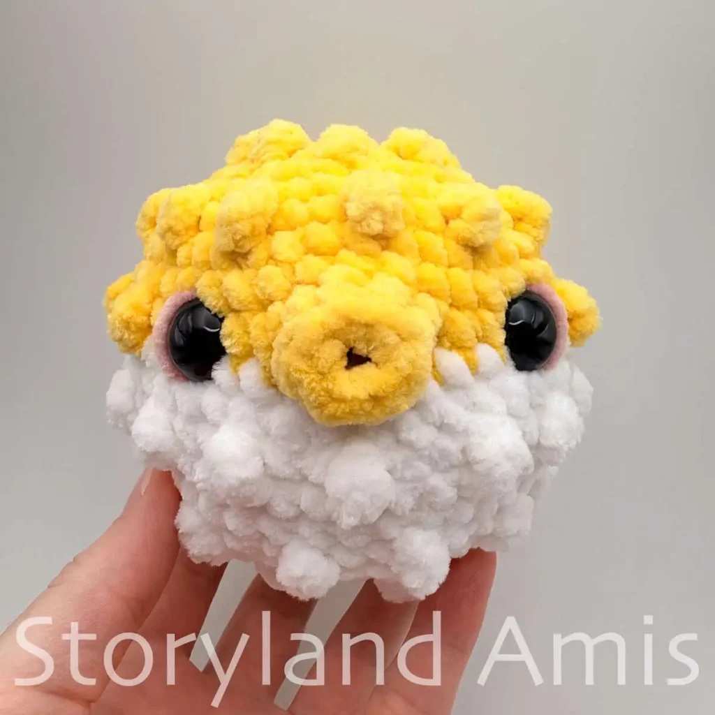 A hand holds up a partially finished white and yellow amigurumi pufferfish toy.