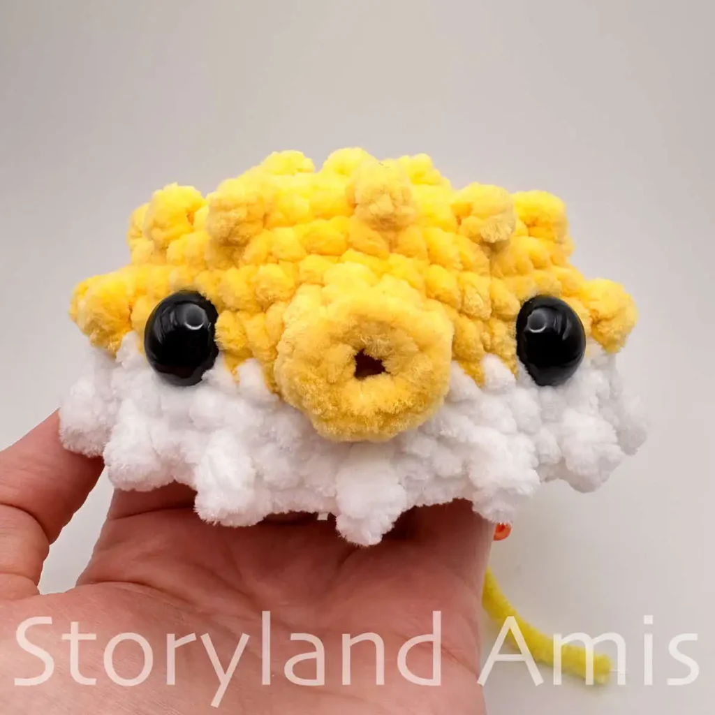 A hand holds a partially finished pufferfish toy with black plastic safety eyes.