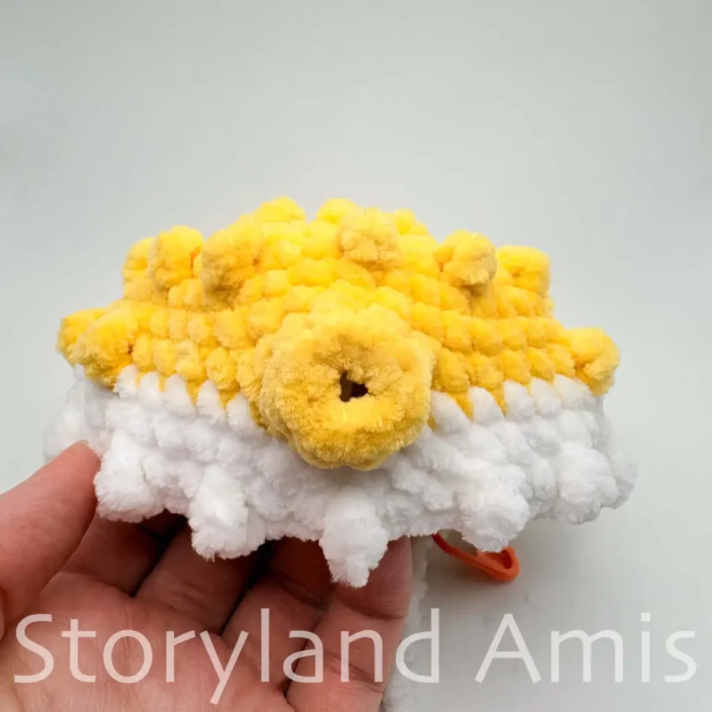 A hand holds a partially finished white and yellow amigurumi pufferfish toy.