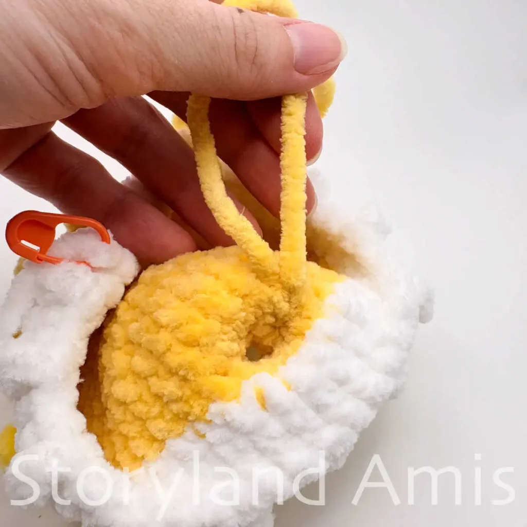 A close-up of a hand holding two yellow yarn tails attached to the inside of an amigurumi pufferfish head.
