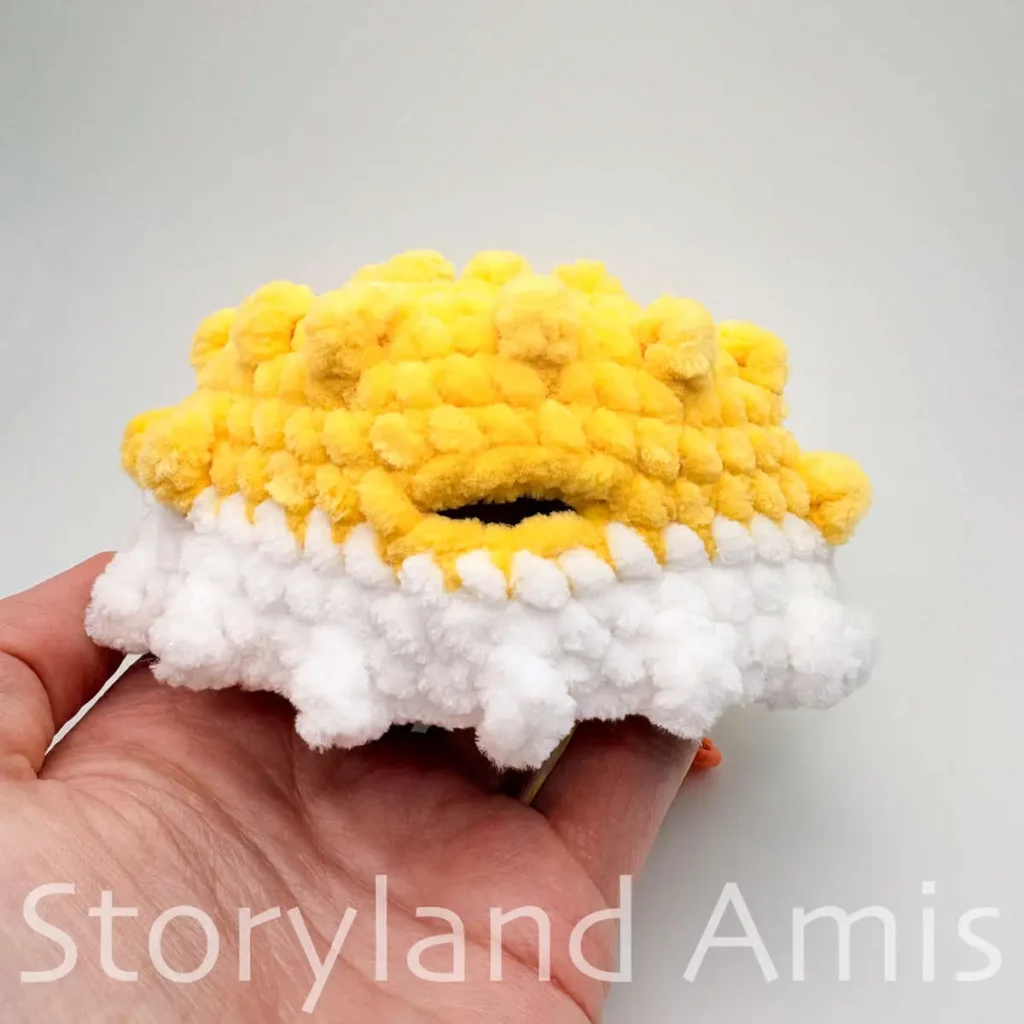 A hand holds a partially finished white and yellow pufferfish amigurumi toy.