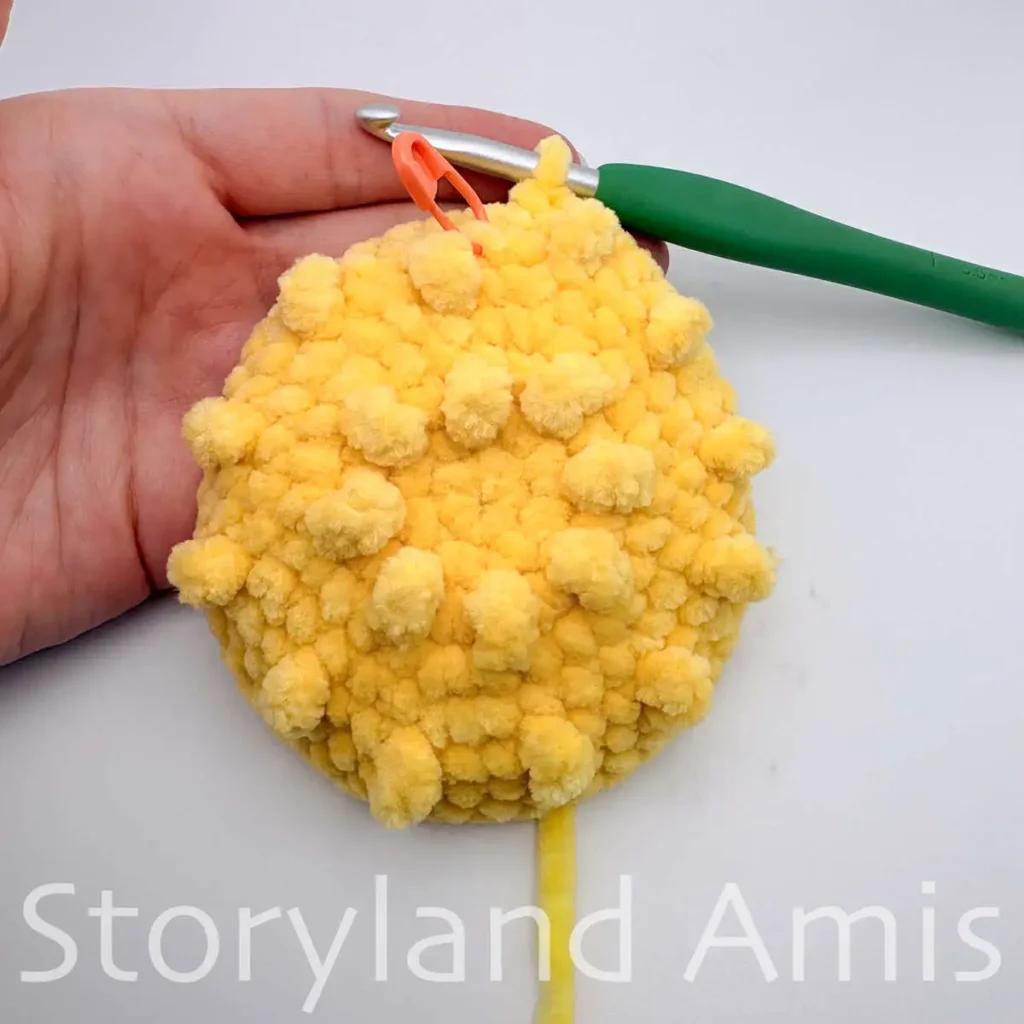 A hand holds a yellow crocheted circle with a green crochet hook inserted in one of the stitches.