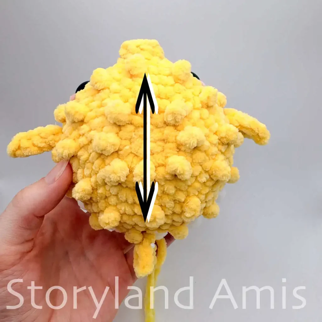 A crocheted amigurumi pufferfish is shown from the top. A black arrow runs parallel over the center back of the pufferfish.